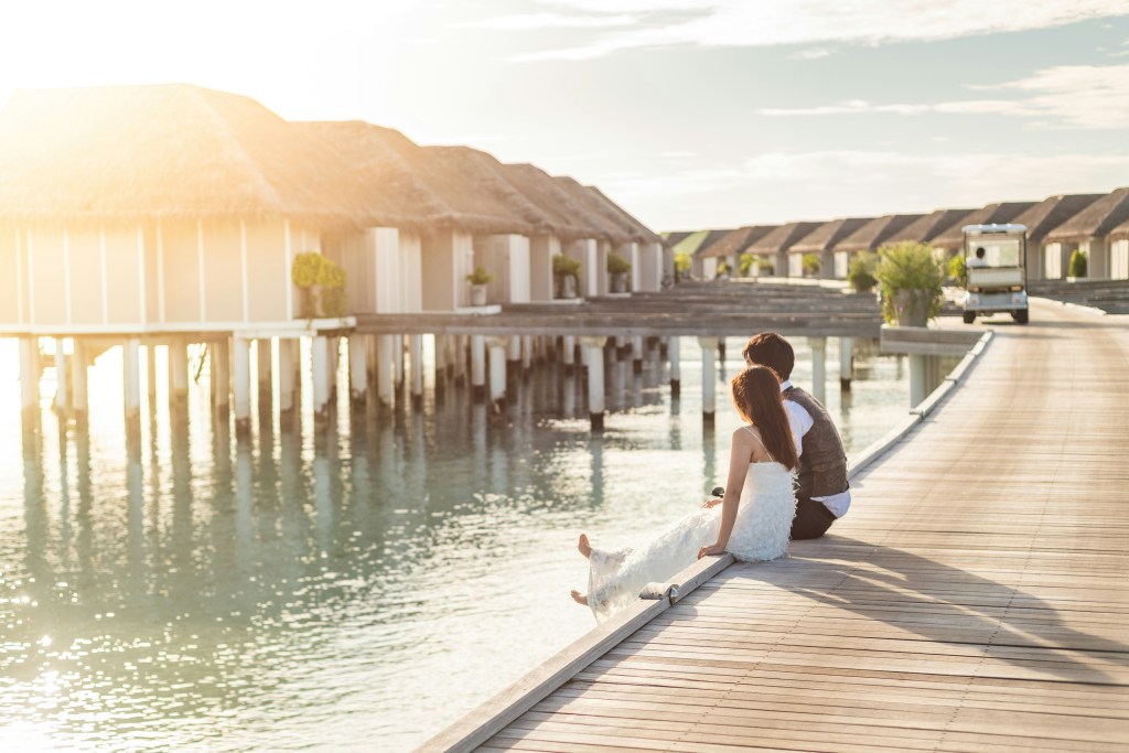 A couple sits on a wooden pier overlooking calm waters at sunset, with overwater bungalows in the background.