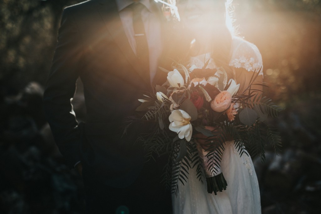 A bride holding a bouquet of flowers stands beside a groom in a suit, with sunlight creating a warm, soft glow around them.