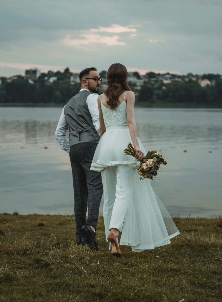A bride in a white gown holds a bouquet while walking along the shore with her groom in a grey vest and checked trousers. The couple is walking away from the camera, with a calm lake and a cloudy sky in the background.