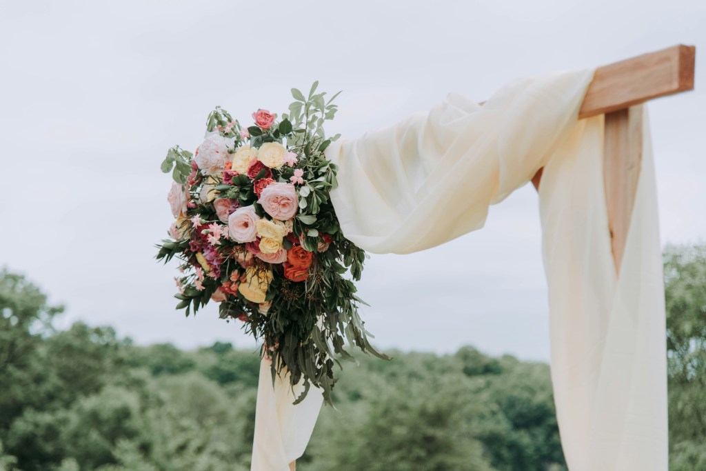 A beautifully arranged bouquet of colourful flowers hanging from a wooden structure draped with soft fabric, set against a natural landscape.