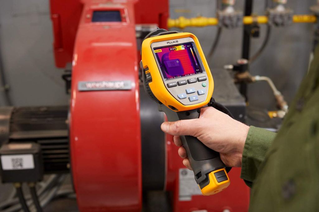 A person holding a Fluke thermal camera, pointing it at a machinery setup with visible temperature readings displayed on the camera screen.