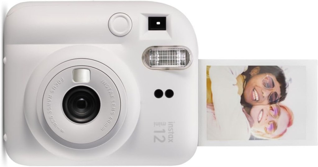 A white instant camera with a round lens and a small screen, producing a printed photo of two smiling individuals wearing sunglasses.