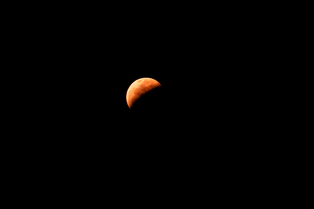 A close-up view of a partially eclipsed moon, showcasing its reddish hue against a dark background.