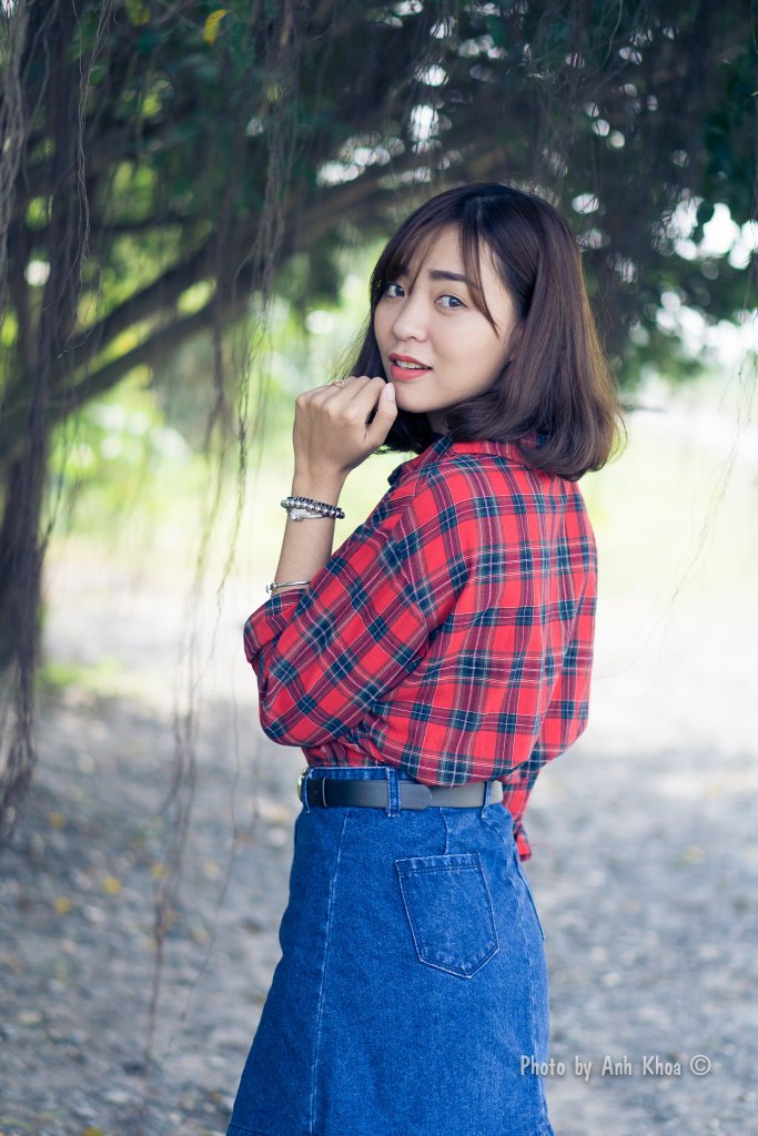 A young woman in a plaid shirt and denim skirt is posing outdoors, looking back over her shoulder with a playful expression. Soft natural lighting filters through the trees in the background.