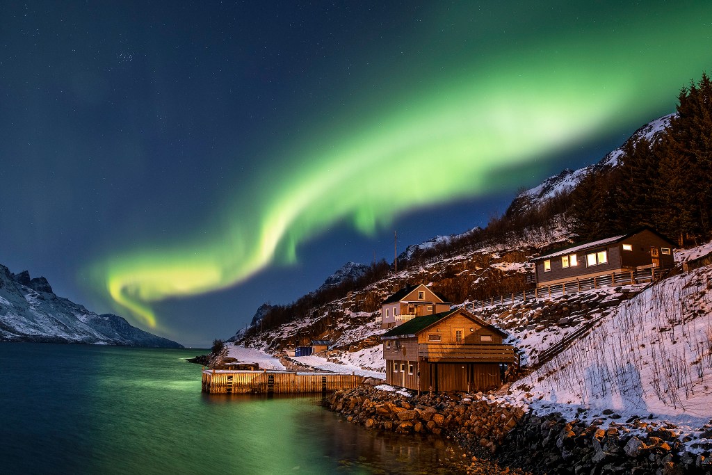A stunning view of the Northern Lights illuminating the night sky over a fjord, with green auroras swirling above snow-covered mountains and wooden houses along the water's edge.