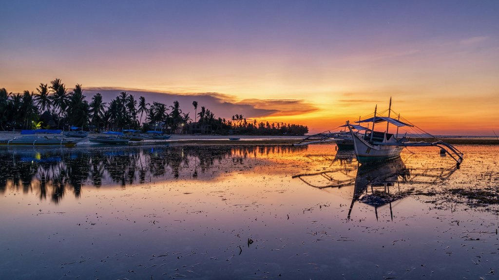 A serene sunset over a calm sea, with fishing boats reflecting on the water and silhouetted palm trees in the background.