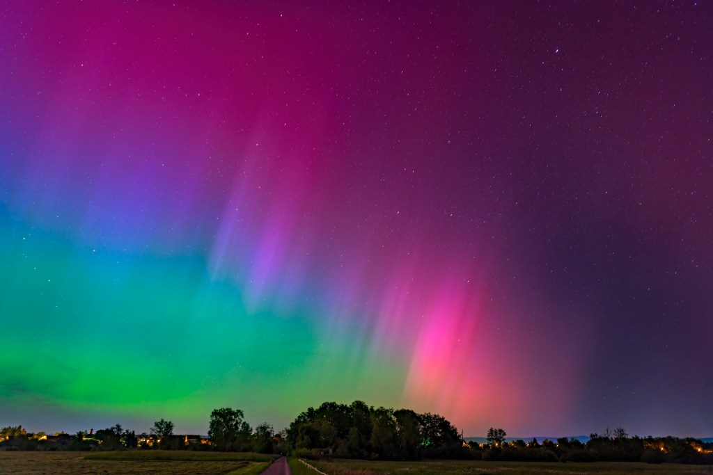 A vibrant display of the Northern Lights featuring shades of green, pink, and purple against a starry night sky, with a tree line silhouette at the bottom and a faint path leading into the scene.