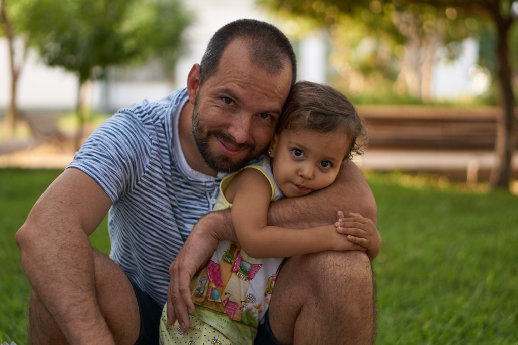 A man smiles while holding a young child in a grassy park setting, both enjoying a moment together.