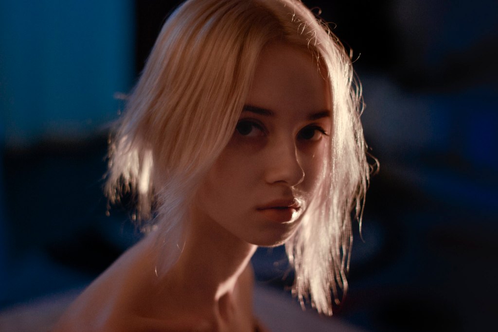 A close-up portrait of a person with short, light-colored hair, featuring soft lighting that highlights the facial features and creates a moody atmosphere.