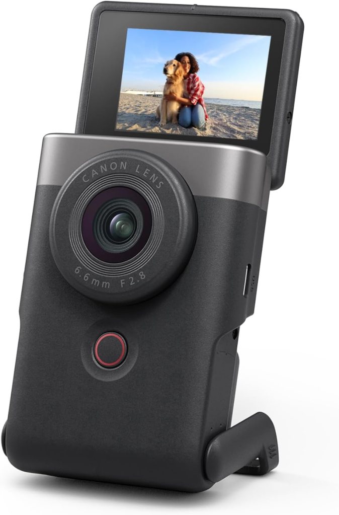 A compact Canon camera with a flip screen displaying a photo of a woman sitting on the beach with a golden retriever.