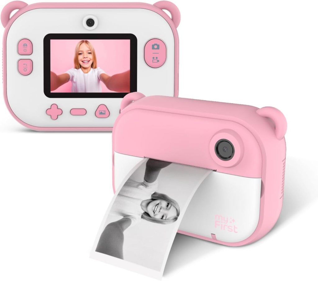 A playful pink children's camera and printer combo featuring an LCD screen displaying a selfie. The camera includes buttons for easy use and a printer is shown producing a black and white photo.